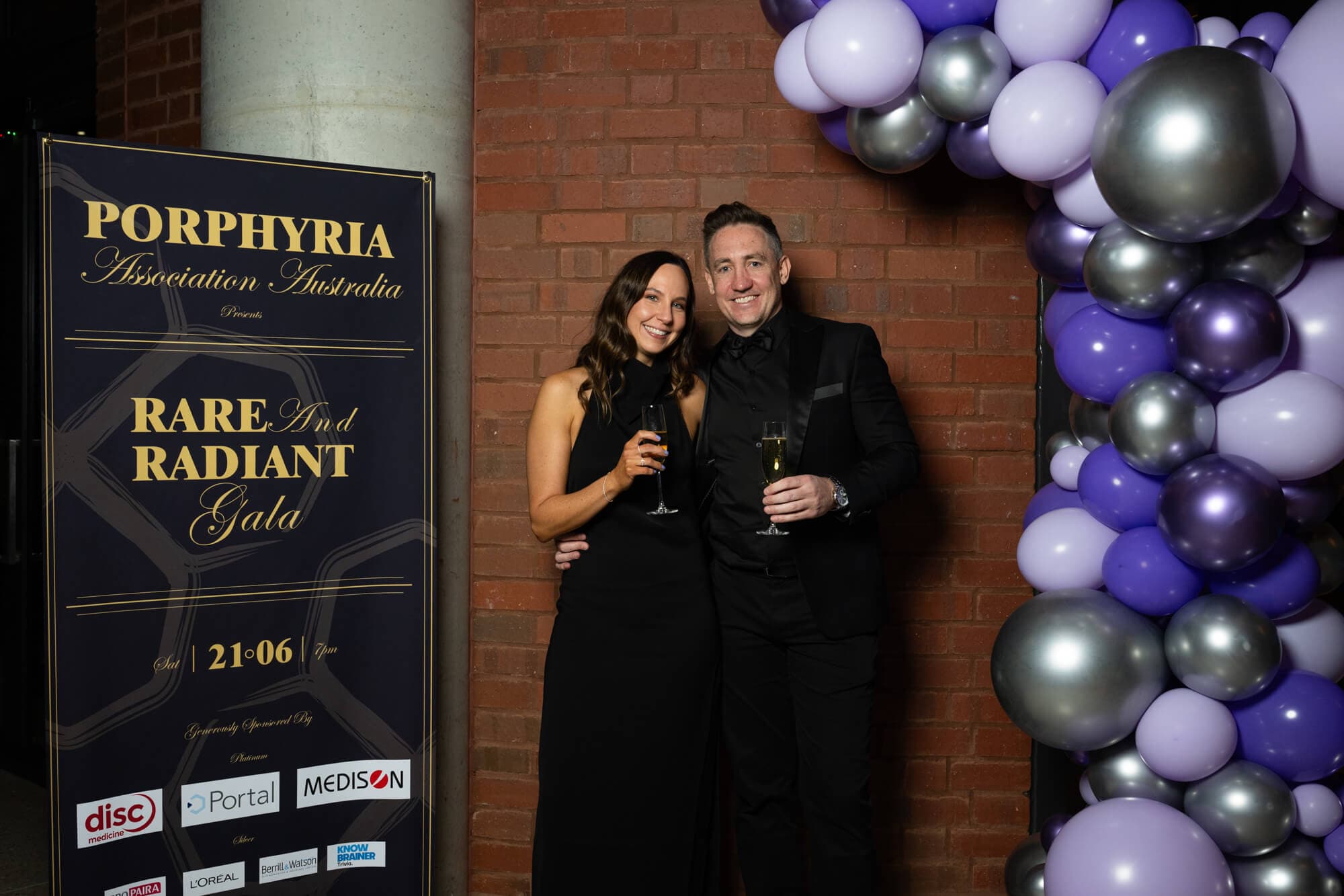 A couple smiling at the media wall beside the purple and silver balloon arch