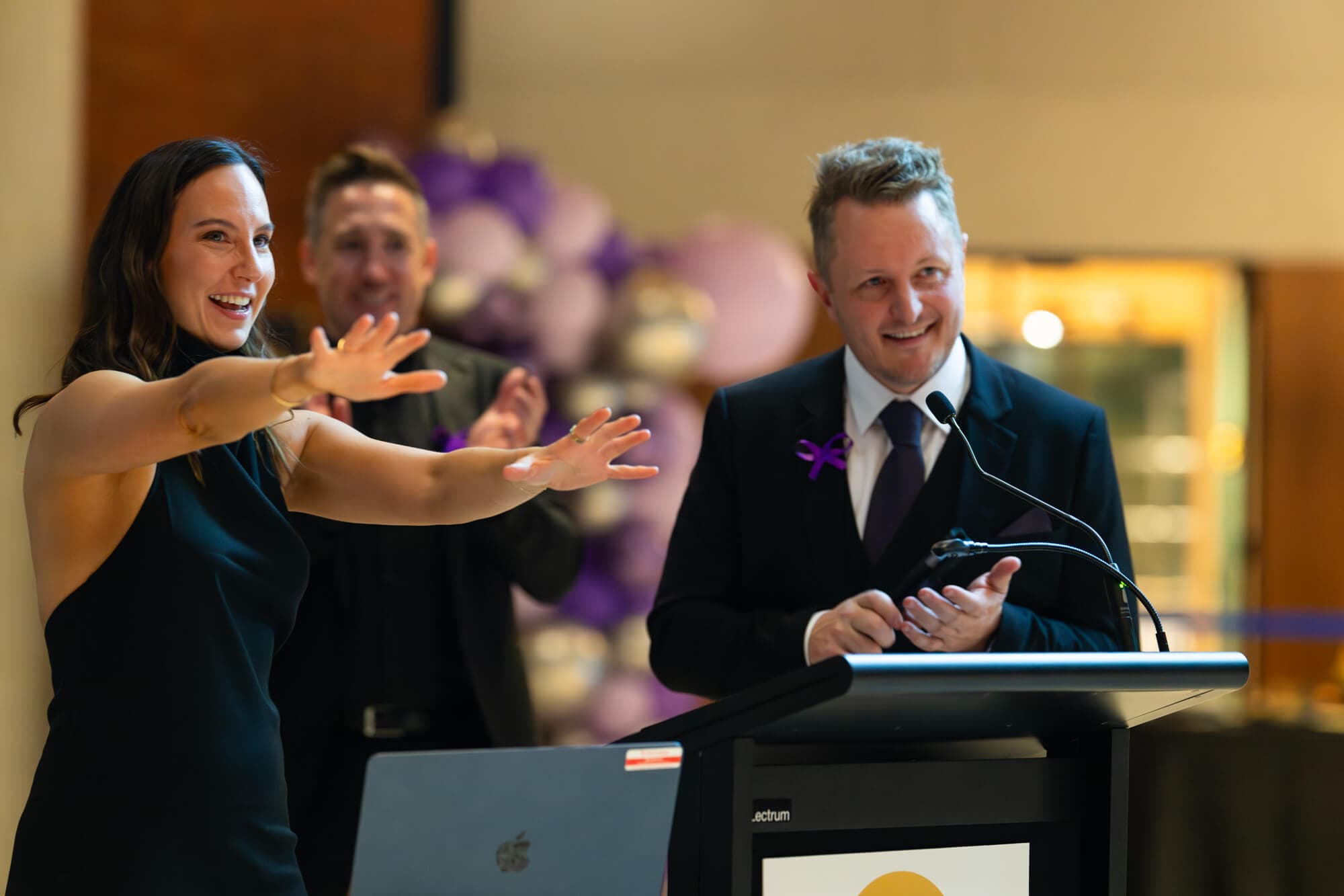 The hosts at the lectern, arms wide, mid-laugh